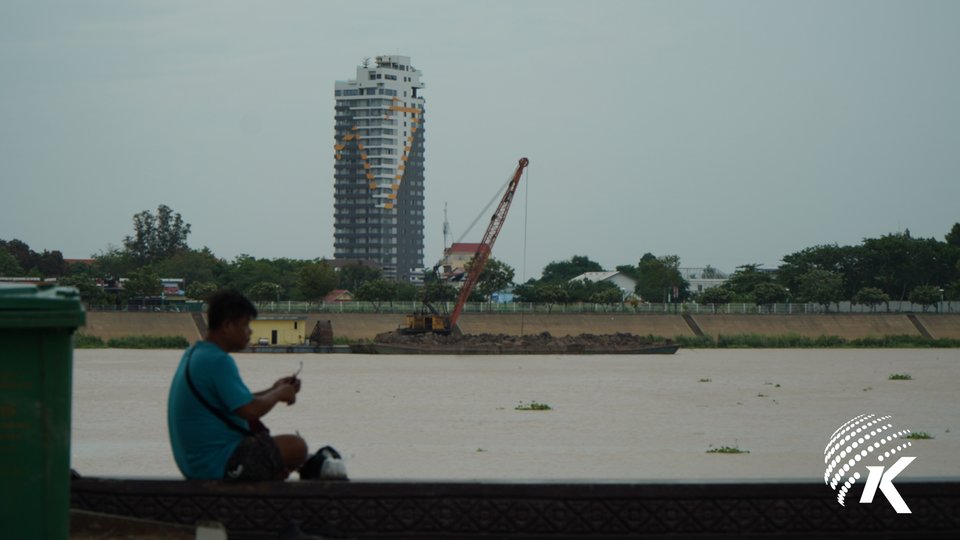 A person sits at the riverside, watching a sand mining ship on the other side. Kiripost/Seng Mengheng